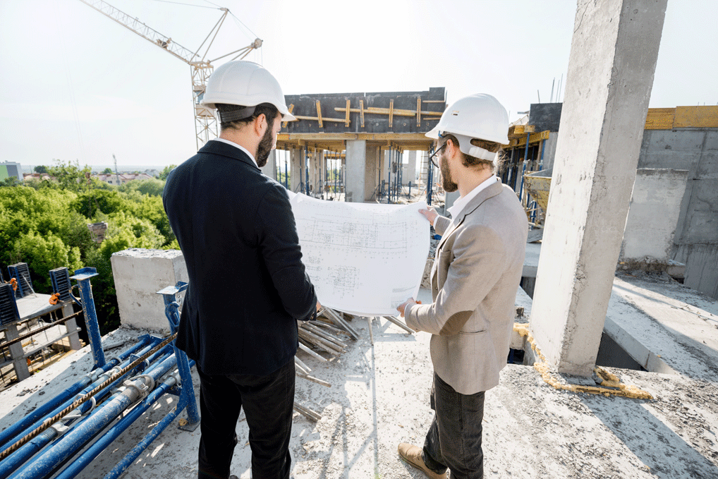 Zwei Bauingenieure mit Schutzhelmen stehen auf einer Baustelle und besprechen gemeinsam Baupläne vor dem Rohbau eines Gebäudes, im Hintergrund ist ein Kran sichtbar.