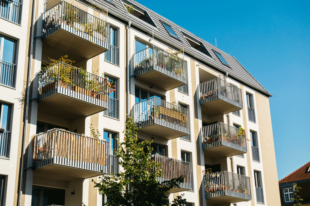 Ein modernes Mehrfamilienhaus mit mehreren Balkonen, die mit Pflanzen geschmückt sind. Die Sonne scheint auf die Fassade des Gebäudes und der Himmel ist klar und blau.