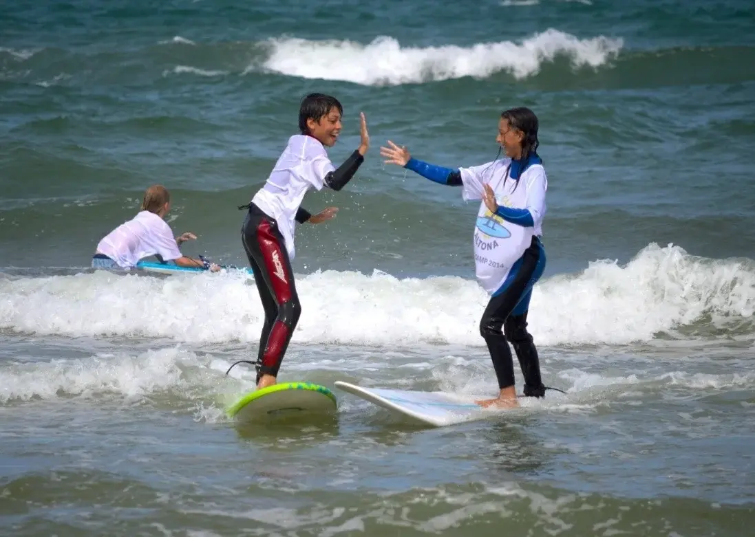 Zwei Kinder in Neoprenanzügen geben sich ein High Five im Wasser während eines Surfkurses