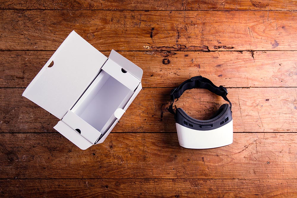 Schwarz weiße VR-Brille neben weißem Verpackungskarton auf einem Holztisch (von oben fotografiert).