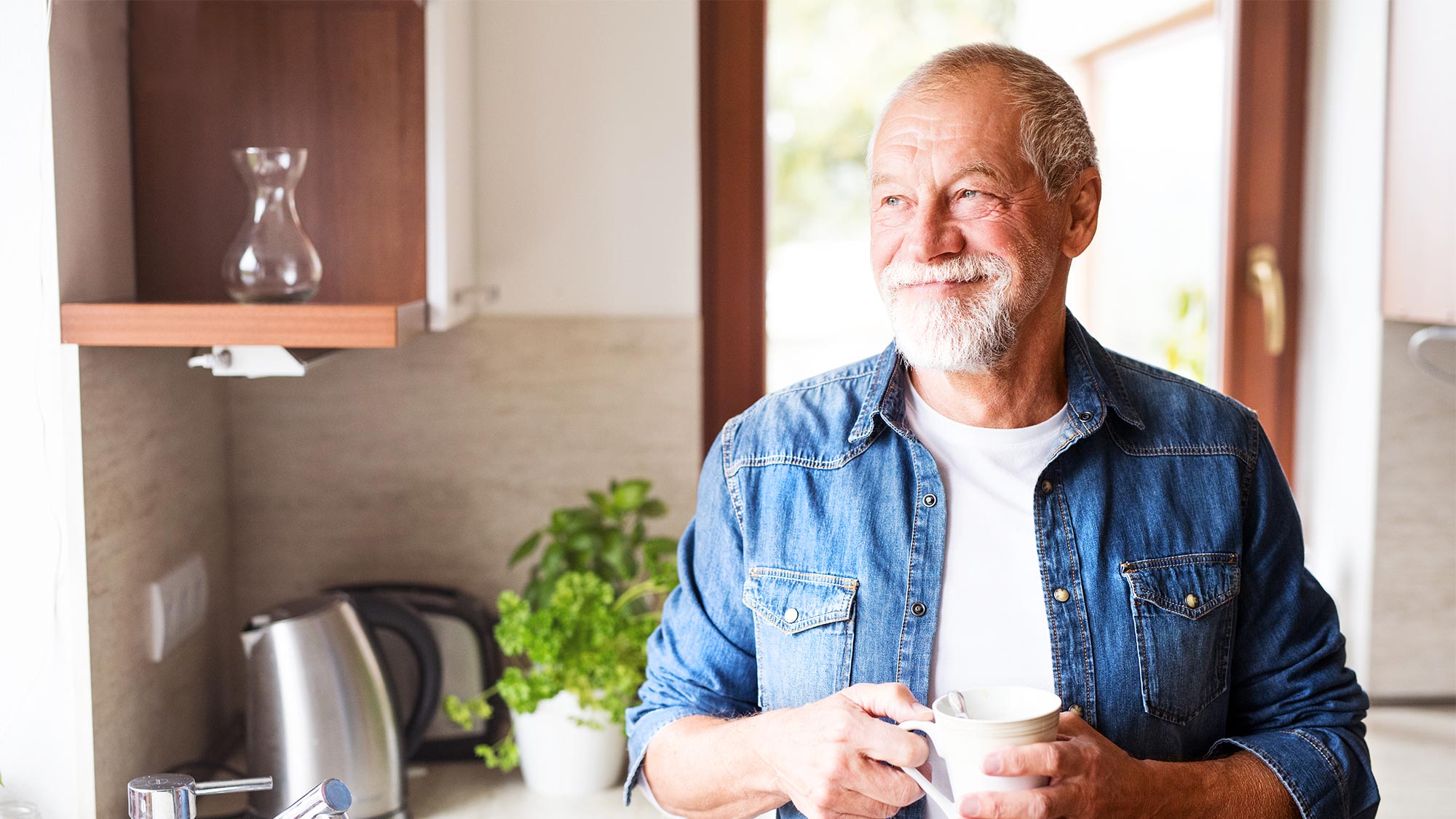 Glücklicher älterer Mann der in seiner Küche steht, aus dem Fenster guckt und eine Tasse Kaffee in der Hand hat.