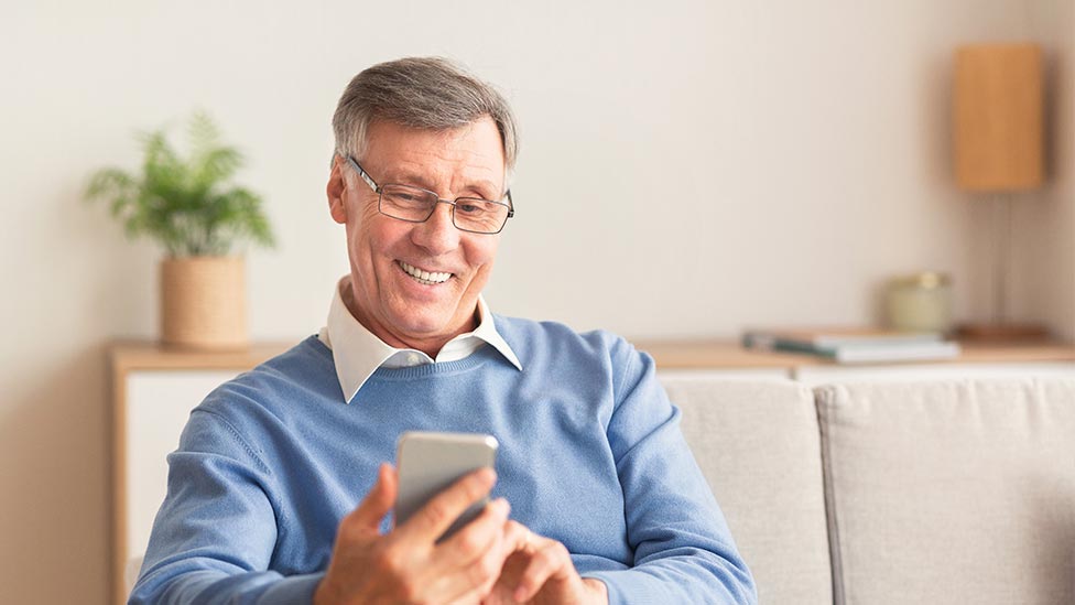 Fröhlicher älterer Mann mit Brille, der im Wohnzimmer auf seiner Couch sitzt und auf sein Smartphone schaut.