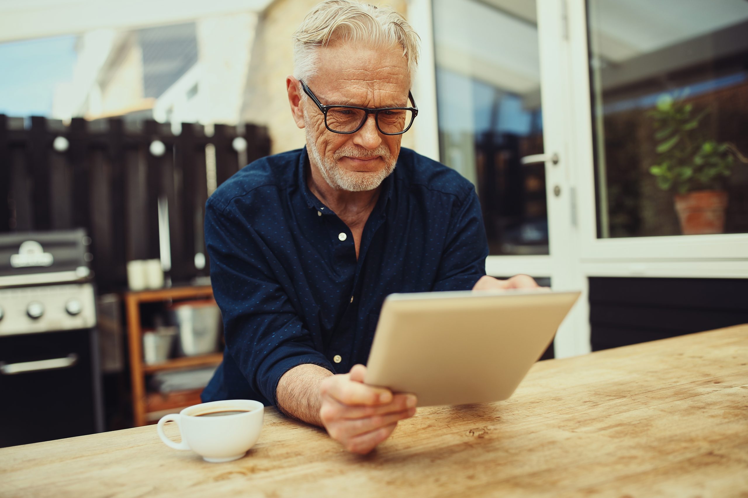 Älterer Herr mit weißen Haar und Brille, der draußen auf seiner Terrasse sitzt und an seinem Tablet Arbeit und Kaffee trinkt.